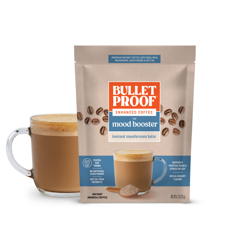 A stand-up pouch of Bulletproof Mood Booster instant mushroom latte coffee stands in front of a glass mug filled with brewed coffee on a white background.