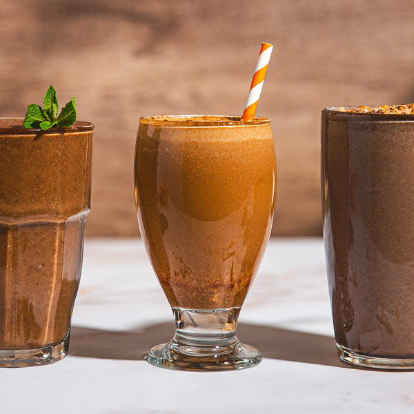 Three chocolate milkshakes in glass cups with a straw on a light surface.