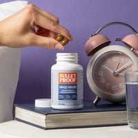 Hand taking a supplement from a Bulletproof 'sleep mode' bottle with an alarm clock and glass of water in the background.
