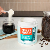 Bulletproof innerfuel prebiotic powder being added to a cup of coffee with coffee beans and a jar in the background.