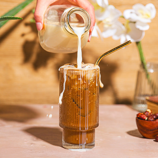 Person pouring a creamy liquid into a glass of iced coffee on a wooden table with flowers in the background.