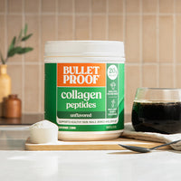 Container of Bulletproof collagen peptides on a kitchen counter with a glass of coffee and a spoon.